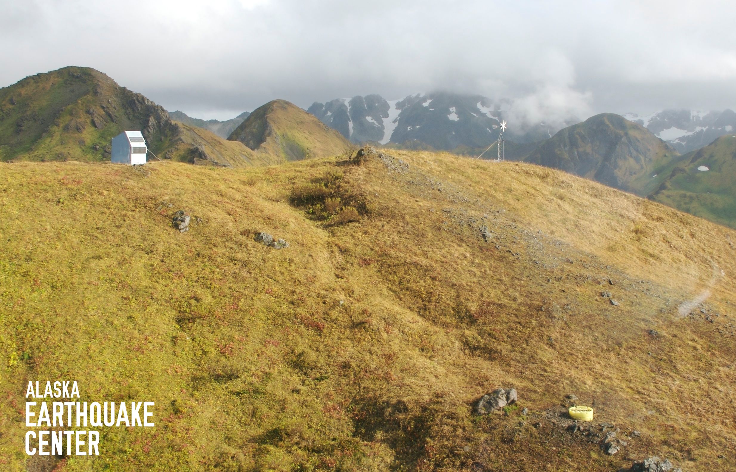 Mountain hillside with seismic station hut at top left and yellow vault for seismometers at bottom right. 