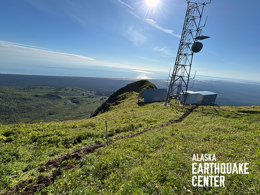 On green bluff is a communications tower with metal sheds next to it in middle ground, with gray cable leading into foreground. Ocean in background.