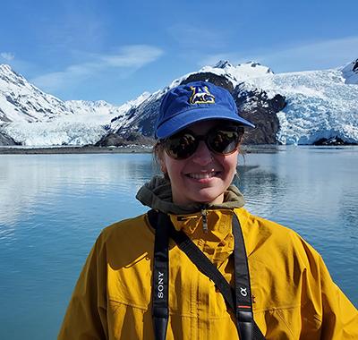 Woman with had and sunglasses with calm ocean and two glaciers in background.