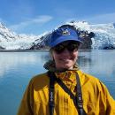 Woman with had and sunglasses with calm ocean and two glaciers in background.